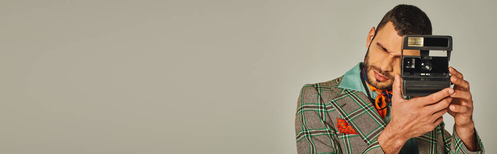 homme à la mode en veste à carreaux et foulard lumineux prenant des photos sur caméra vintage sur gris, bannière - Photo, image