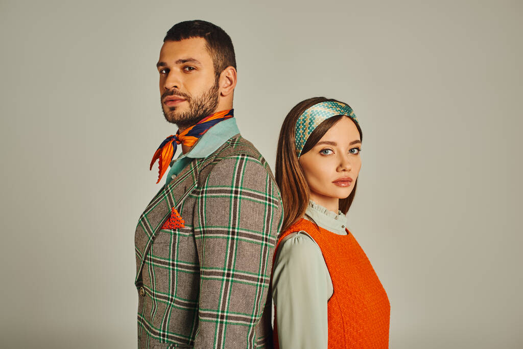 young couple in stylish old-fashioned attire standing back to back and looking at camera on grey - Photo, Image