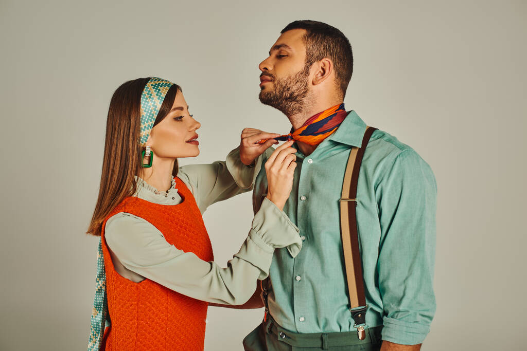 очаровательная женщина привязывает яркий шейный платок к стильному мужчине в подтяжках на серой, винтажной моде - Фото, изображение