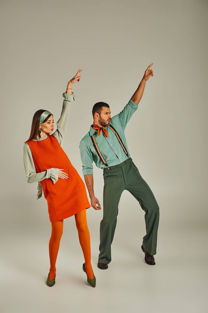 stijlvol paar in vintage kleding dansen met opgeheven handen op grijs, retro vibes en opwinding - Foto, afbeelding