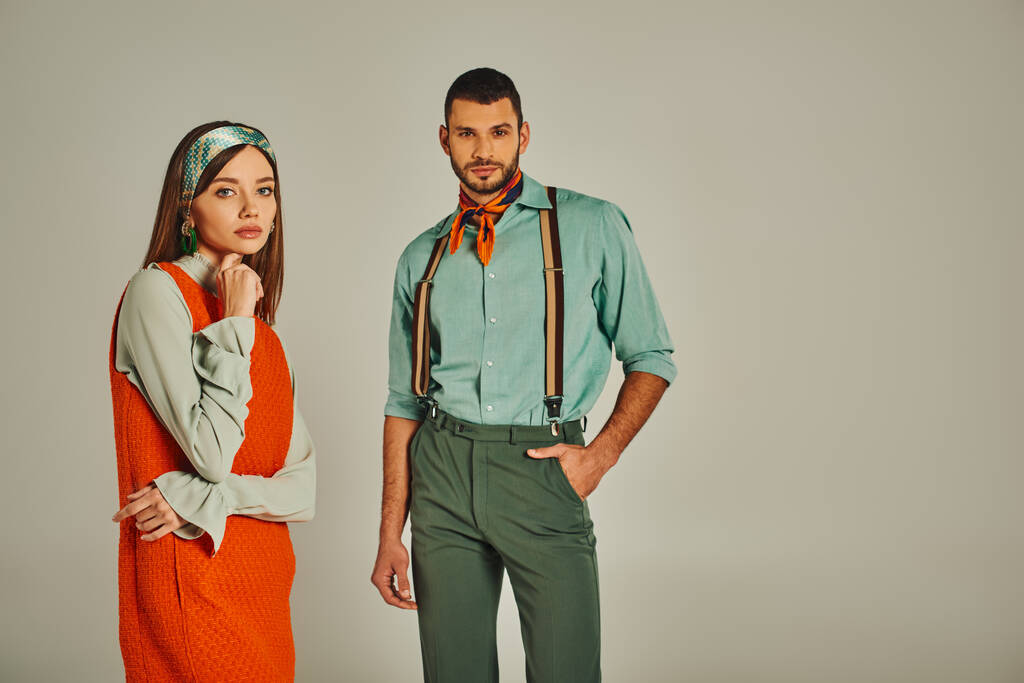 confident man with hand in pocket near thoughtful woman in orange dress on grey, retro fashion - Photo, Image