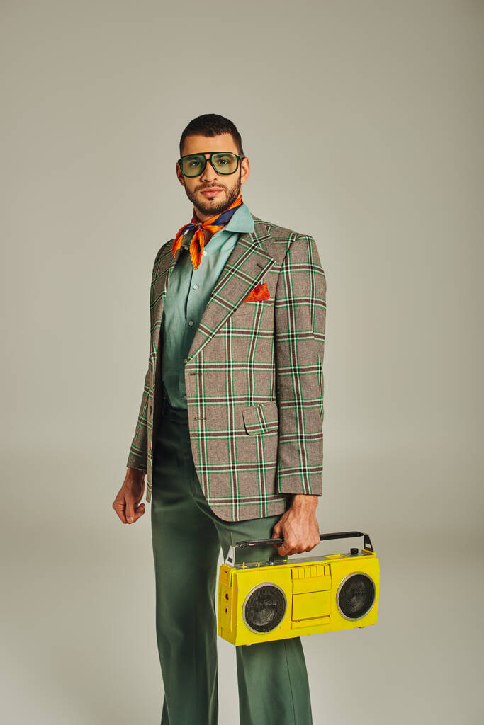 Trendiger Mann in kariertem Blazer und Sonnenbrille mit gelber Boombox auf grauem, retro-inspiriertem Lifestyle - Foto, Bild