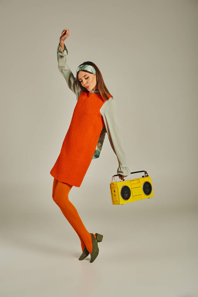 vzrušená žena v oranžových šatech tančící se žlutým boomboxem a zvednutou rukou na šedé, retro vibrace - Fotografie, Obrázek