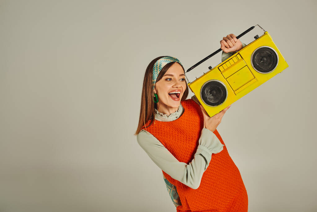 overjoyed woman in orange retro style dress holding yellow boombox and dancing on grey, happiness - Photo, Image