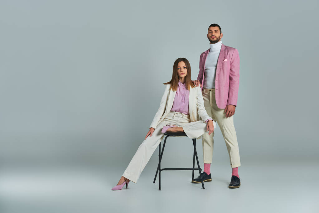 elegant woman in white suit posing on chair near trendy man in lilac blazer on grey, full length - Photo, Image