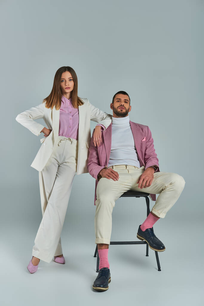 woman in white formal suit posing with hand on hip near man in lilac blazer sitting on chair on grey - Photo, Image
