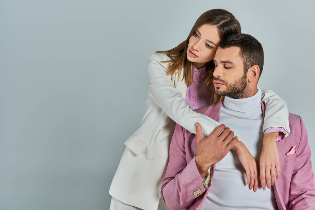 mujer joven en traje blanco abrazando al hombre en blazer lila en gris, pareja con elegante atuendo formal - Foto, imagen