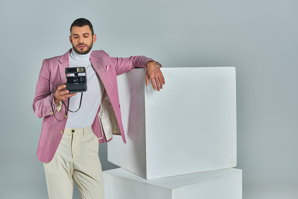 молодой человек в стильной формальной одежде позирует рядом с белыми кубиками с винтажной камерой на серой, современной моде - Фото, изображение