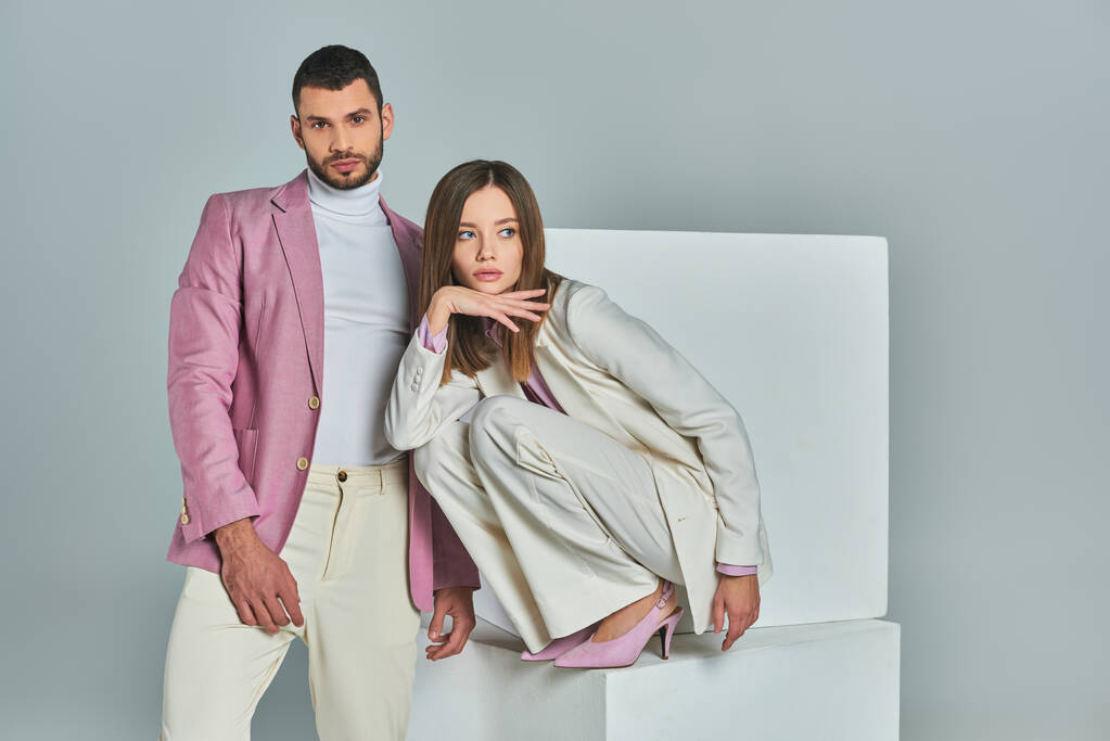 confident man in lilac blazer looking at camera near stylish woman posing on white cubes on grey - Photo, Image