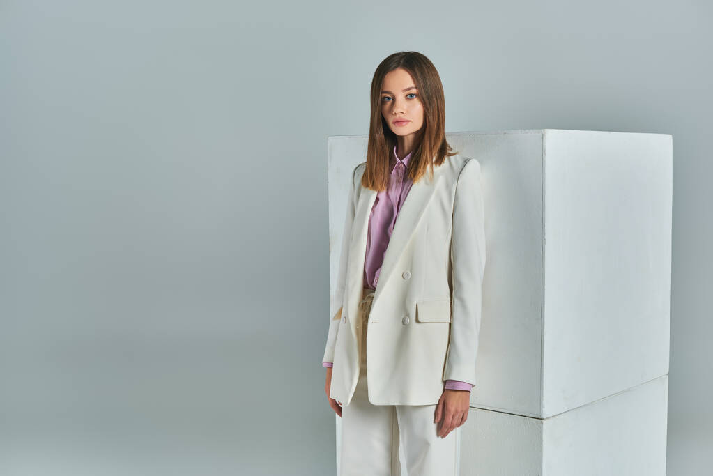 minimalistic fashion, young woman in stylish formal wear looking at camera near white cubes on grey - Photo, Image