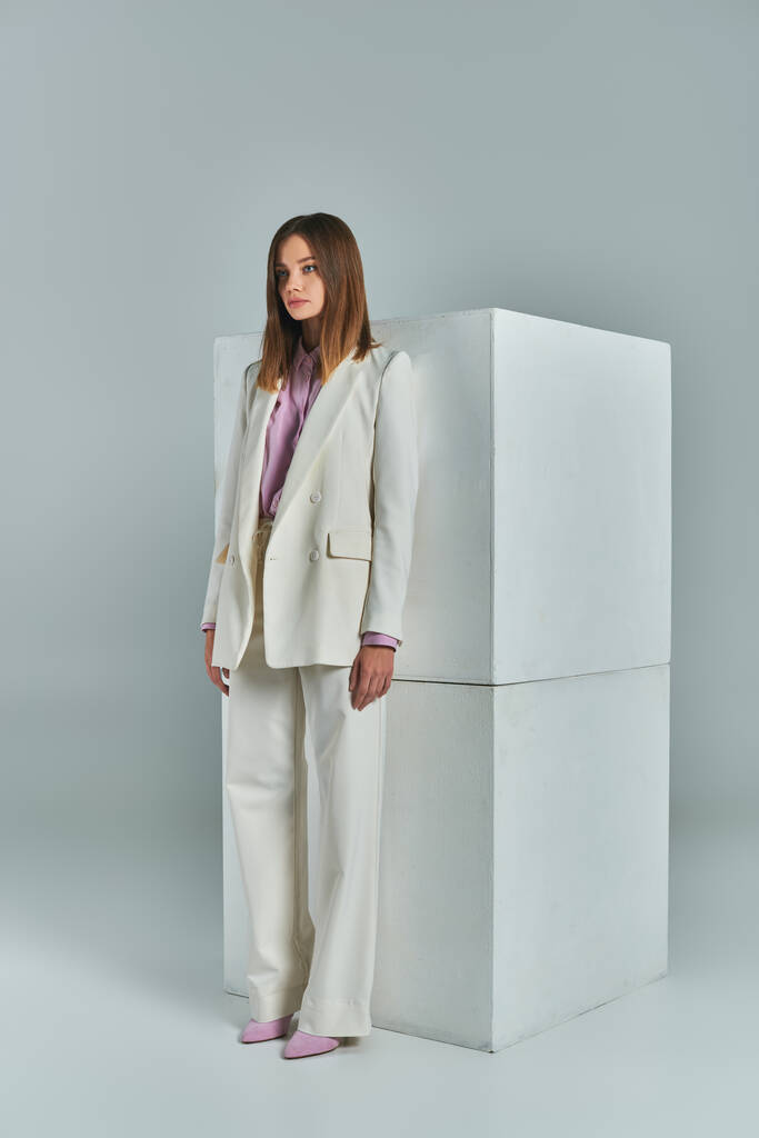 traje de negocios de moda, mujer moderna en traje elegante blanco de pie cerca de cubos en gris, longitud completa - Foto, imagen