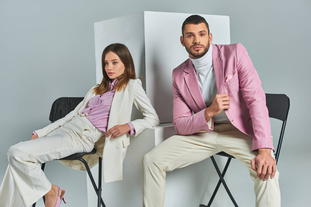 νεαρό ζευγάρι σε μοντέρνα επίσημη φθορά κάθεται σε καρέκλες κοντά σε κύβους σε γκρι, μοντέρνα επιχειρηματική μόδα - Φωτογραφία, εικόνα