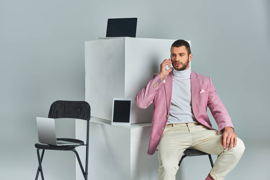 уверенный стильный мужчина разговаривает на смартфоне, сидя рядом с устройствами на белых кубиках на сером - Фото, изображение