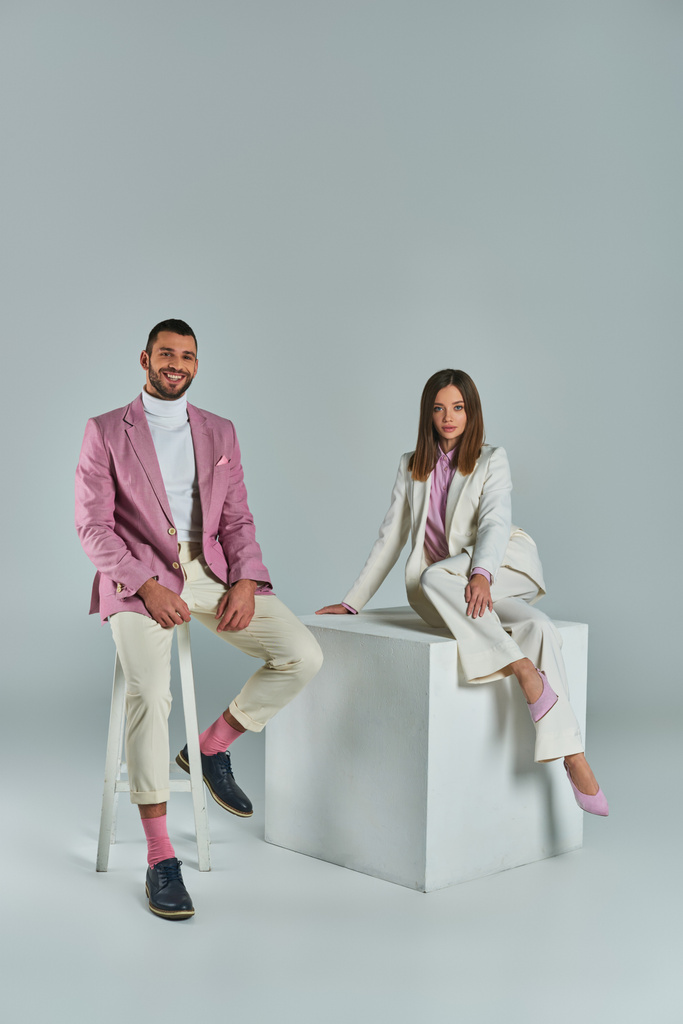 homme élégant souriant sur chaise près de la femme en costume posant sur cube blanc sur gris, mode d'affaires - Photo, image