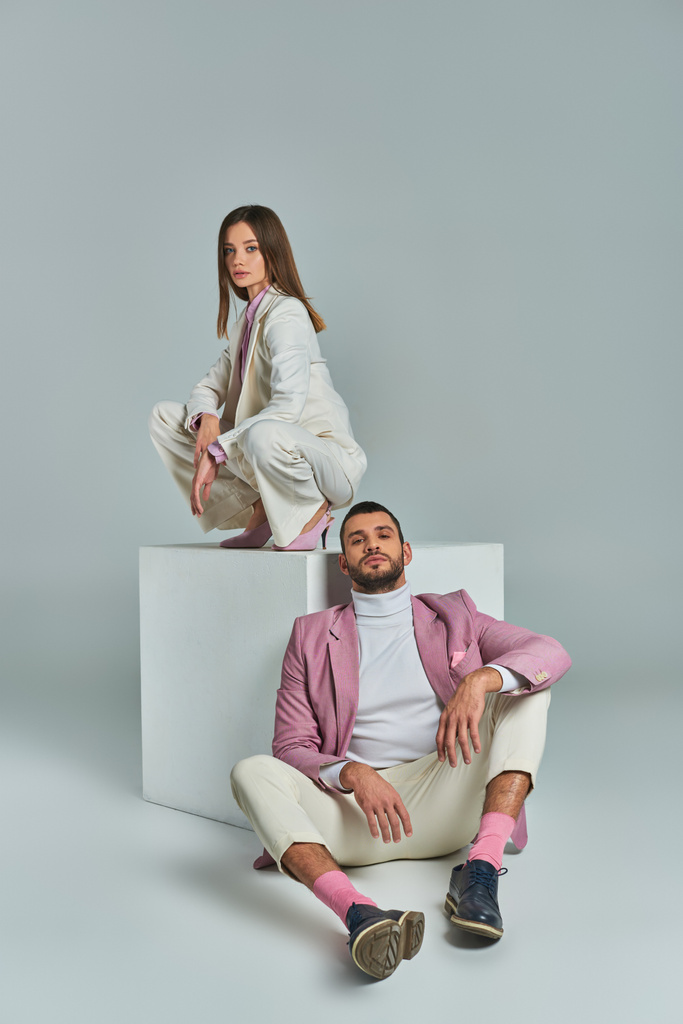 Mann in fliederfarbenem Blazer sitzt auf dem Boden neben Frau im eleganten Anzug und posiert auf weißem Würfel auf grau - Foto, Bild