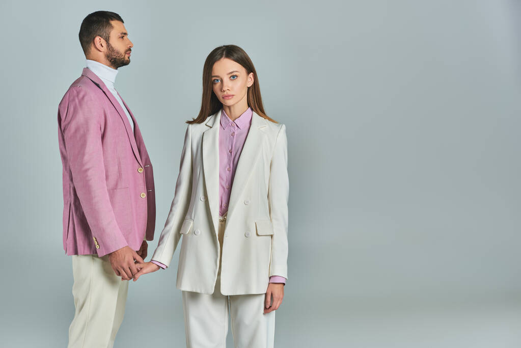 mujer joven en traje blanco cogido de la mano con el hombre en blazer lila y mirando a la cámara en gris - Foto, imagen