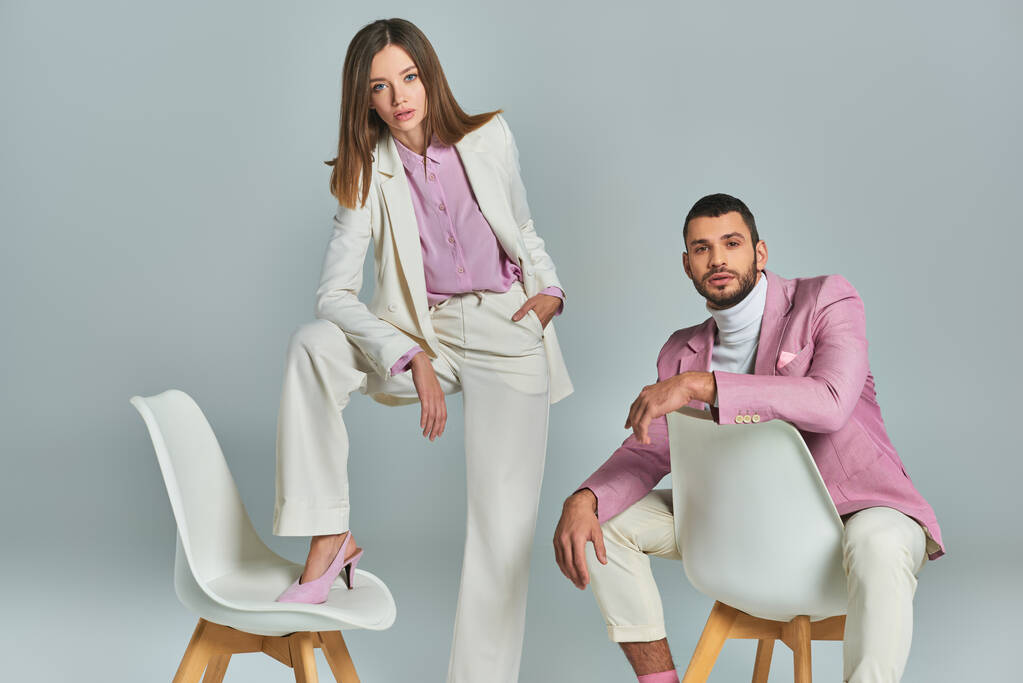 trendiger Mann in fliederfarbenem Blazer sitzt neben Frau im weißen eleganten Anzug und posiert mit Sessel auf grau - Foto, Bild