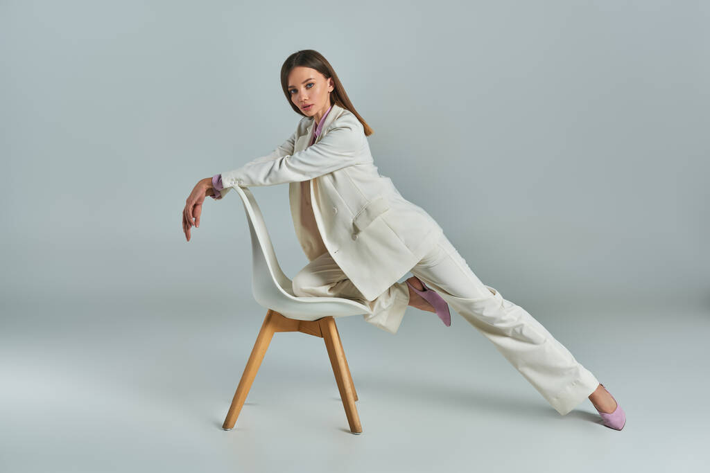 longitud completa de la mujer expresiva en traje blanco posando con sillón y mirando a la cámara en gris - Foto, Imagen