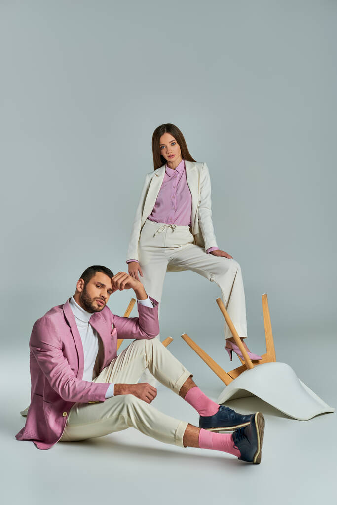 fashionable couple in elegant formal wear posing with overturned armchairs on grey, modern fashion - Photo, Image