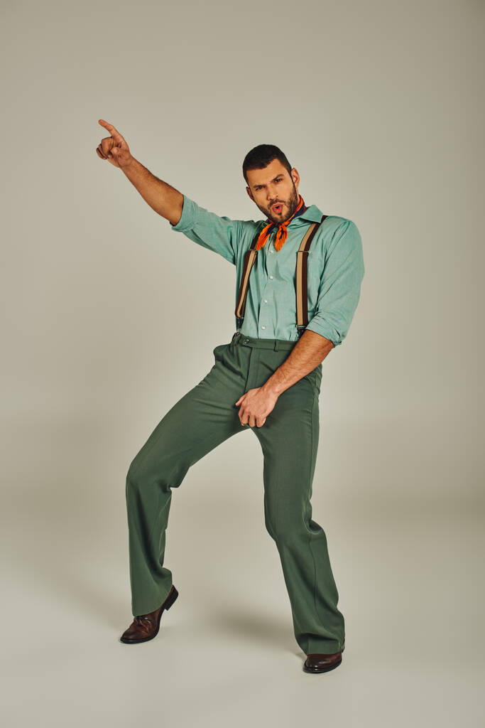aufgeregter Mann in Retro-Klamotten und Hosenträgern, der mit dem Finger zeigt und auf grauen Ganzkörperanzügen tanzt - Foto, Bild