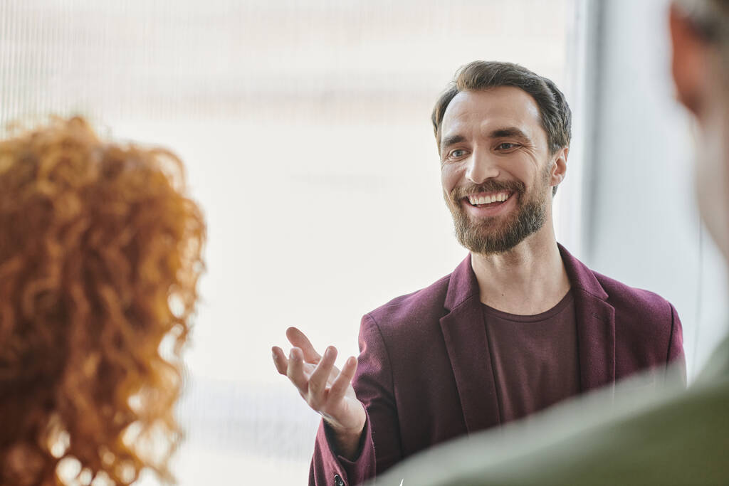 χαμογελαστός γενειοφόρος επιχειρηματίας μιλάει και gesturing κοντά κοκκινομάλλα γυναίκα σε θολή πρώτο πλάνο στο γραφείο - Φωτογραφία, εικόνα