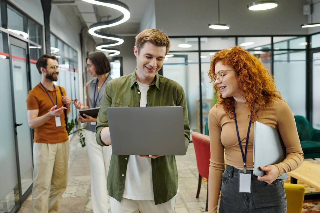 redhead businesswoman pointing at laptop near colleague, coworkers talking on background in office - Photo, Image