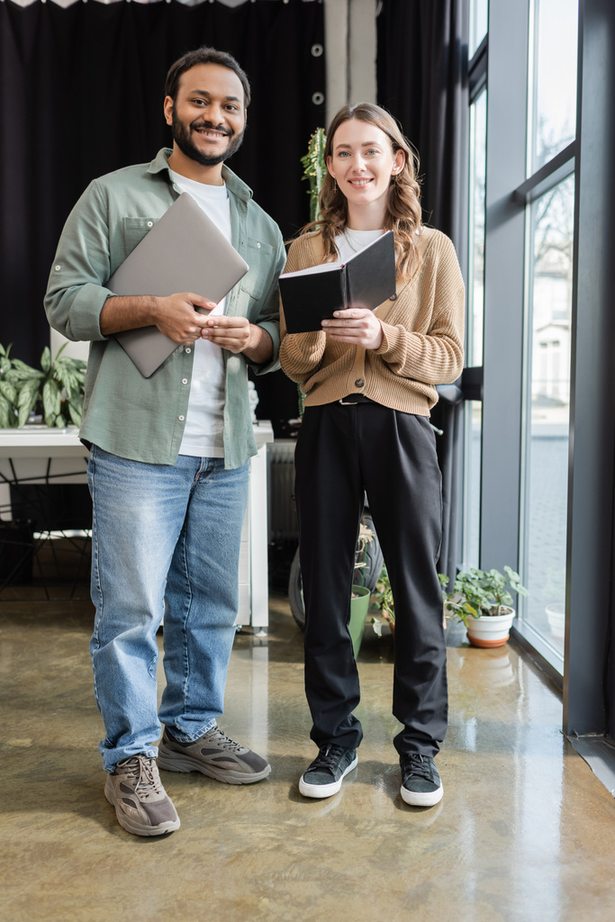 cheerful woman with notebook and indian man with laptop looking at camera in office, coworking space - Photo, Image