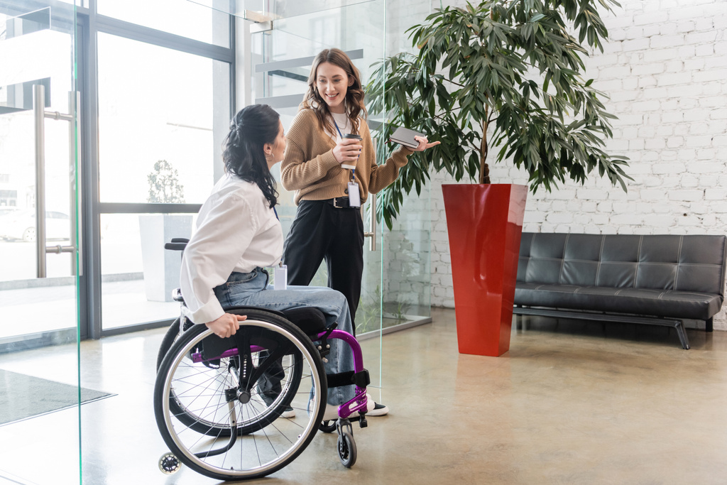 Inklusionskonzept, glückliche Frau zeigt weg und blickt behinderte Kollegin im Rollstuhl an - Foto, Bild