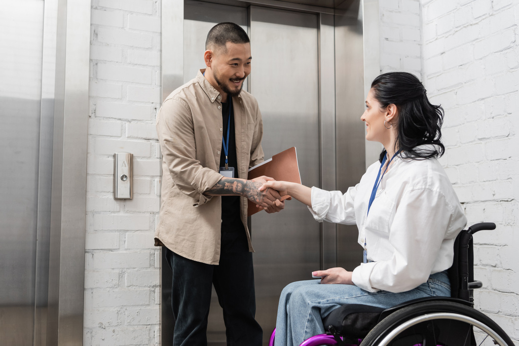 inclusie en diversiteit, vrolijke aziatische man schudden handen met gehandicapte vrouw in de buurt van kantoor liften - Foto, afbeelding
