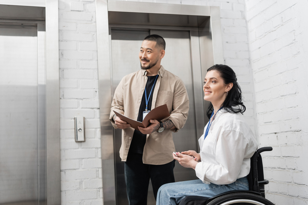 включення і різноманітність, щаслива азіатська папка чоловіка біля інвалідів жінки та офісних ліфтів, дивіться геть - Фото, зображення