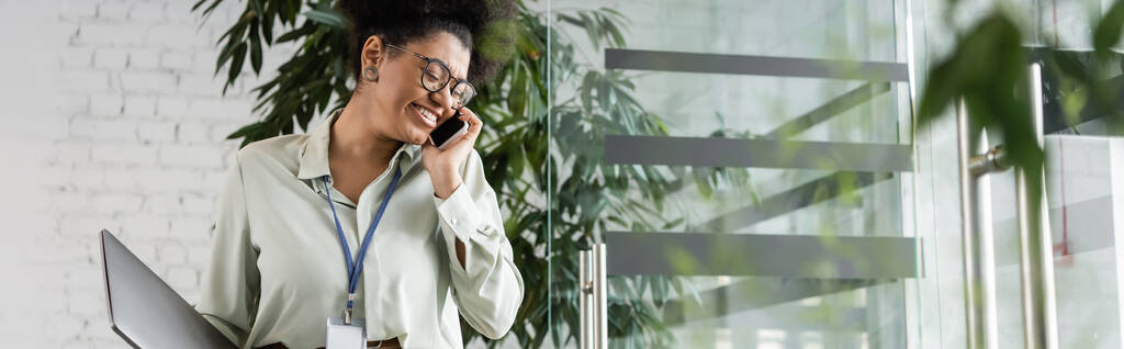 alegre mujer de negocios afroamericana en gafas que sostienen el ordenador portátil y hablan en el teléfono inteligente, bandera - Foto, imagen