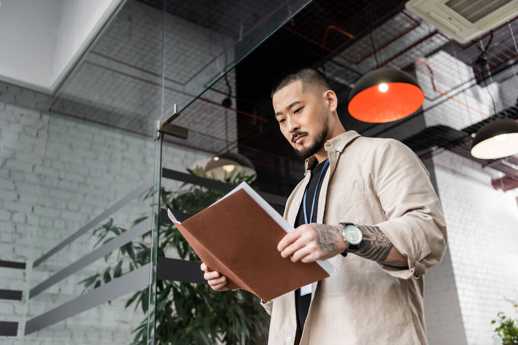 focused asian team leader with tattoo looking at startup plan inside of folder near glass door - Photo, Image