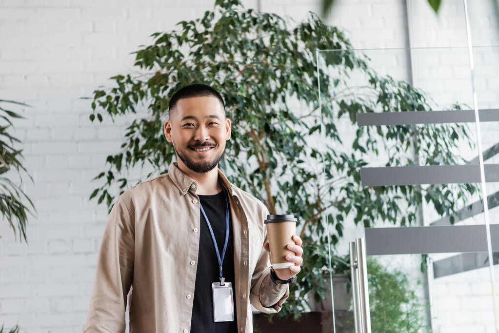 ευτυχισμένος ασιάτης επιχειρηματίας με τατουάζ κρατώντας καφέ για να πάει και κοιτάζοντας κάμερα, εταιρική ζωή - Φωτογραφία, εικόνα