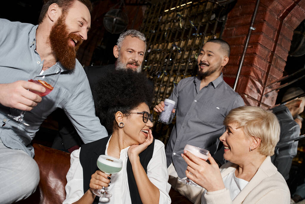 boldog többnemzetiségű nők koktélokkal beszélgetnek szakállas férfiak közelében a bárban, szórakozás és szabadidő munka után - Fotó, kép