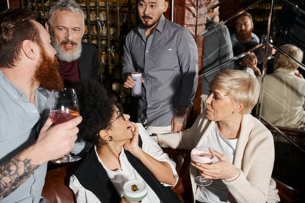 бородаті чоловіки розмовляють біля здивованих жінок, багатоетнічні колеги з коктейлями, що розслабляються в барі - Фото, зображення