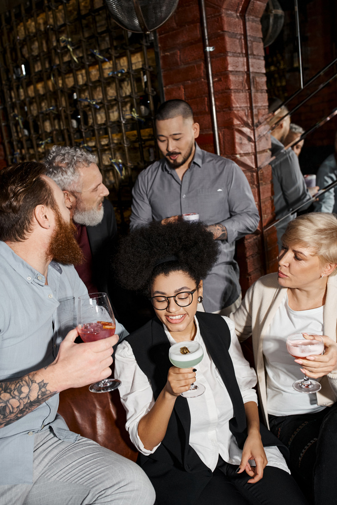 hombres tatuados barbudos hablando cerca de mujeres bebiendo cócteles en el bar, ocio de colegas multiétnicos - Foto, Imagen