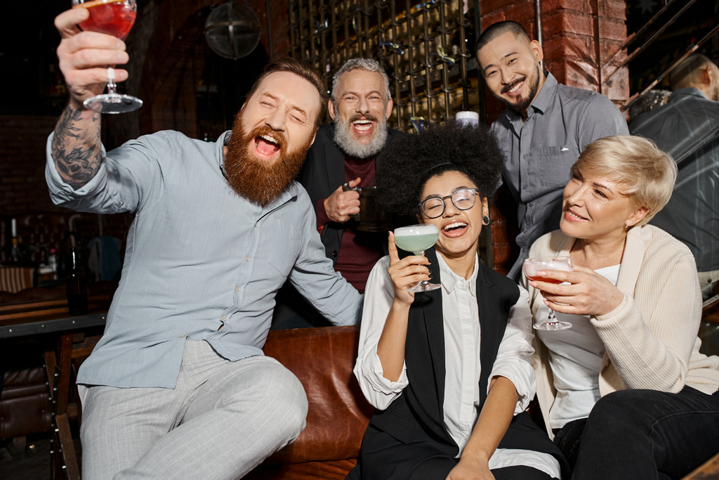 возбужденные многонациональные друзья, держащие напитки и смеющиеся в коктейль-баре, веселящиеся после работы - Фото, изображение