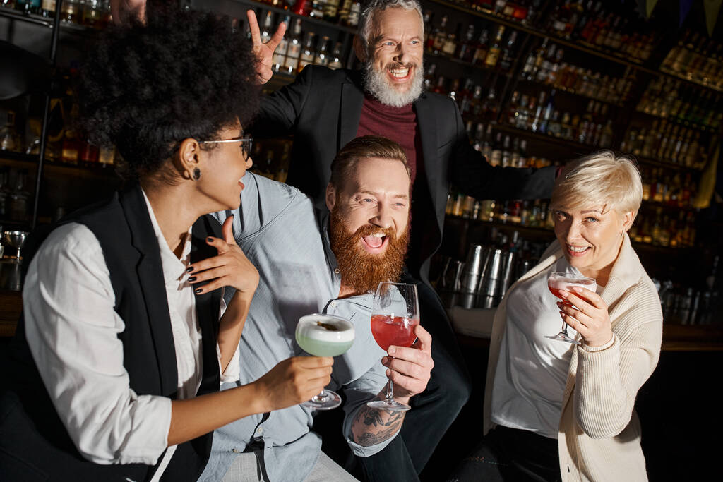 Heyecanlı sakallı adam, barda kokteyl içen çoklu etnik arkadaşların yanında zafer işareti gösteriyor. - Fotoğraf, Görsel