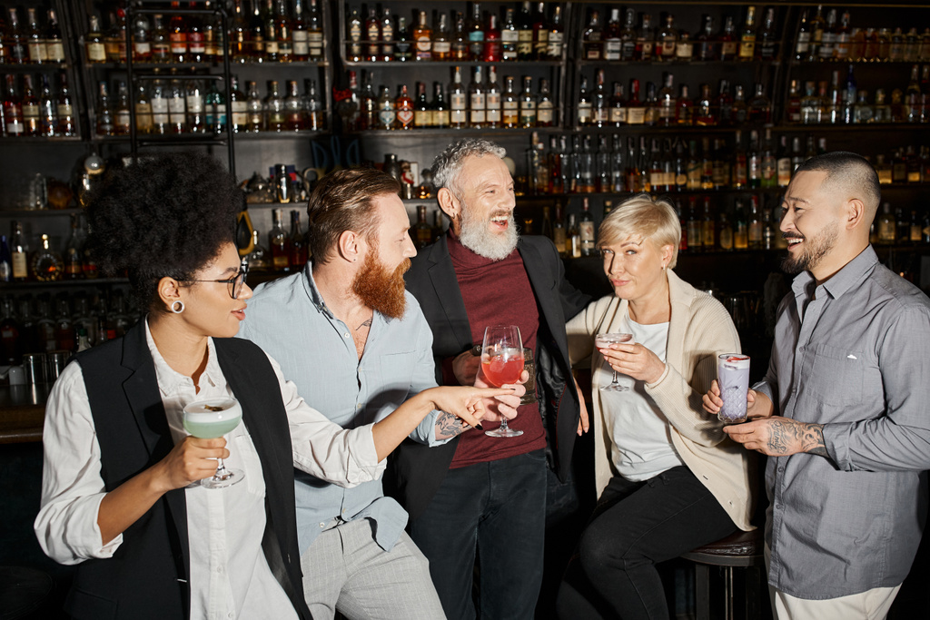 Женщина средних лет, смотрящая в камеру рядом с позитивными мультикультурными друзьями, пьет коктейли в баре - Фото, изображение