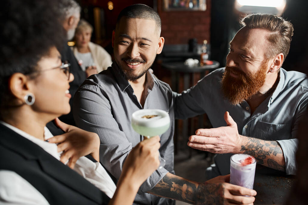 brodaty wytatuowany mężczyzna rozmawia ze zdumioną afrykańską Amerykanką blisko uśmiechnięty azjatycki kolega w barze - Zdjęcie, obraz