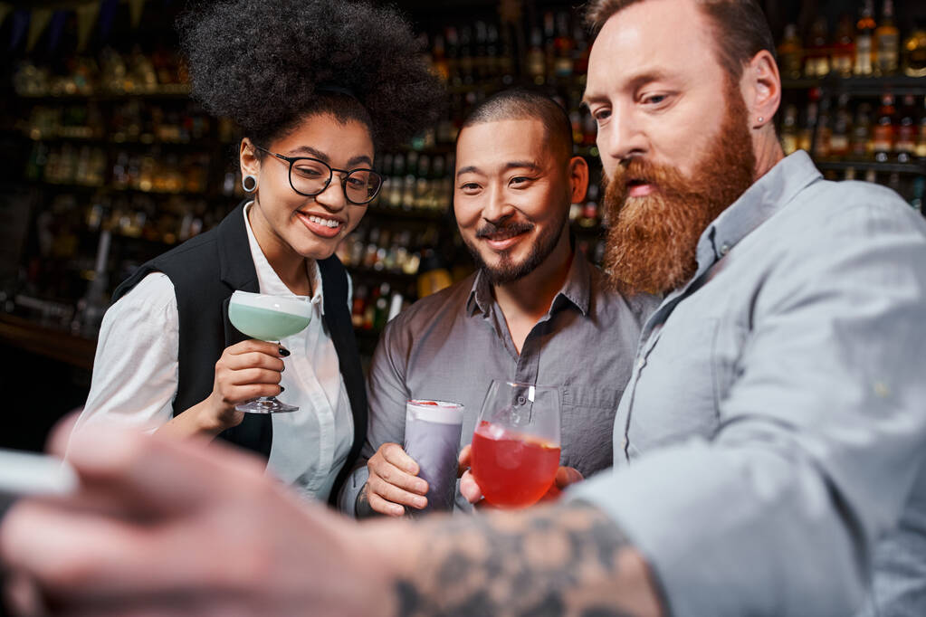 татуированный бородатый мужчина фотографируется на смартфоне с счастливыми мультикультурными коллегами в коктейль-баре - Фото, изображение