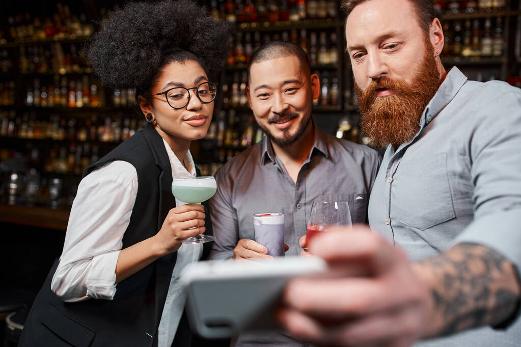 wytatuowany brodaty mężczyzna robi zdjęcie na smartfonie ze szczęśliwymi wieloetnicznymi przyjaciółmi z pracy w barze - Zdjęcie, obraz