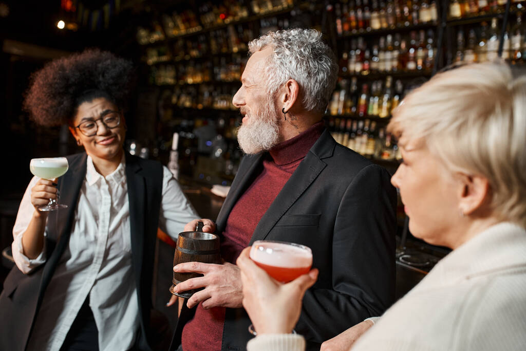 barbuto uomo di mezza età sorridente vicino a donne multietniche con cocktail, colleghi che riposano nel bar - Foto, immagini