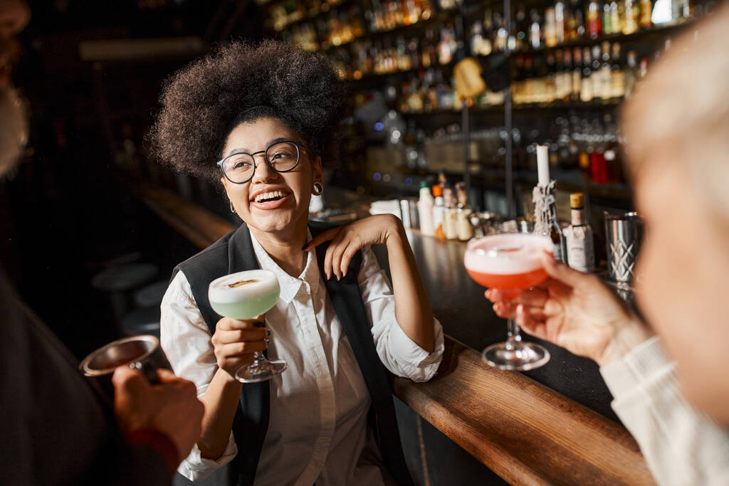 fröhliche afrikanisch-amerikanische Frau hält Cocktailglas in der Hand, während sie Zeit mit Kollegen in der Bar verbringt - Foto, Bild