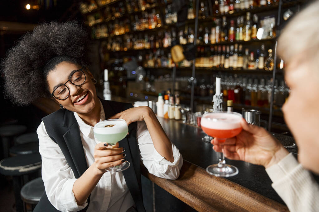 veselý africký Američan žena drží koktejl sklo v blízkosti ženské kolegyně na rozmazané popředí - Fotografie, Obrázek