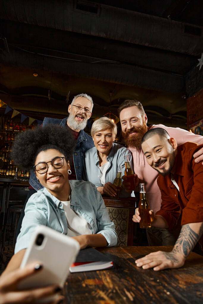 mulher americana africana alegre mostrando sinal de rock e tomando selfie com colegas de trabalho multiétnicos no pub - Foto, Imagem