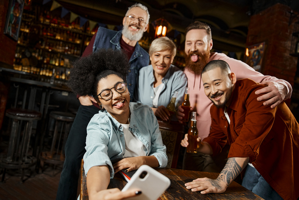 zabawna Afroamerykanka wystająca z języka i robiąca selfie z wielonarodowymi kolegami z pracy w barze - Zdjęcie, obraz