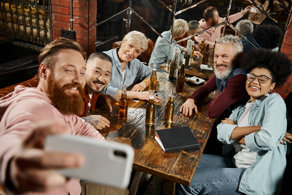 hombre barbudo feliz tomando selfie con colegas multiétnicos cerca de botellas de cerveza en la mesa de madera en el pub - Foto, imagen