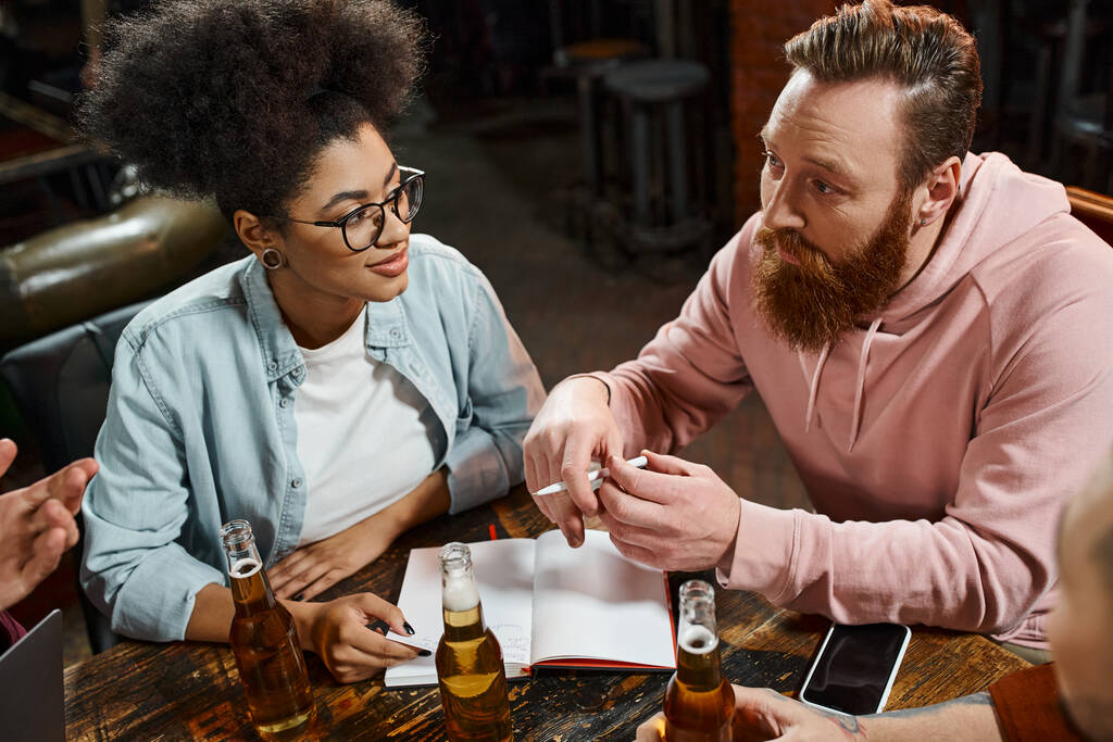 homme barbu parler à l'équipe multiethnique alors qu'il était assis dans un pub près d'un ordinateur portable et bouteilles de bière - Photo, image