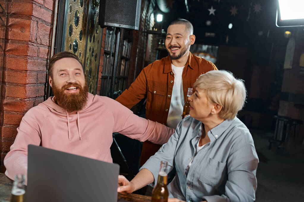 iloinen parrakas mies hymyilee lähellä kannettavaa tietokonetta ja monikansallisia kollegoita puhumassa pubissa töiden jälkeen - Valokuva, kuva